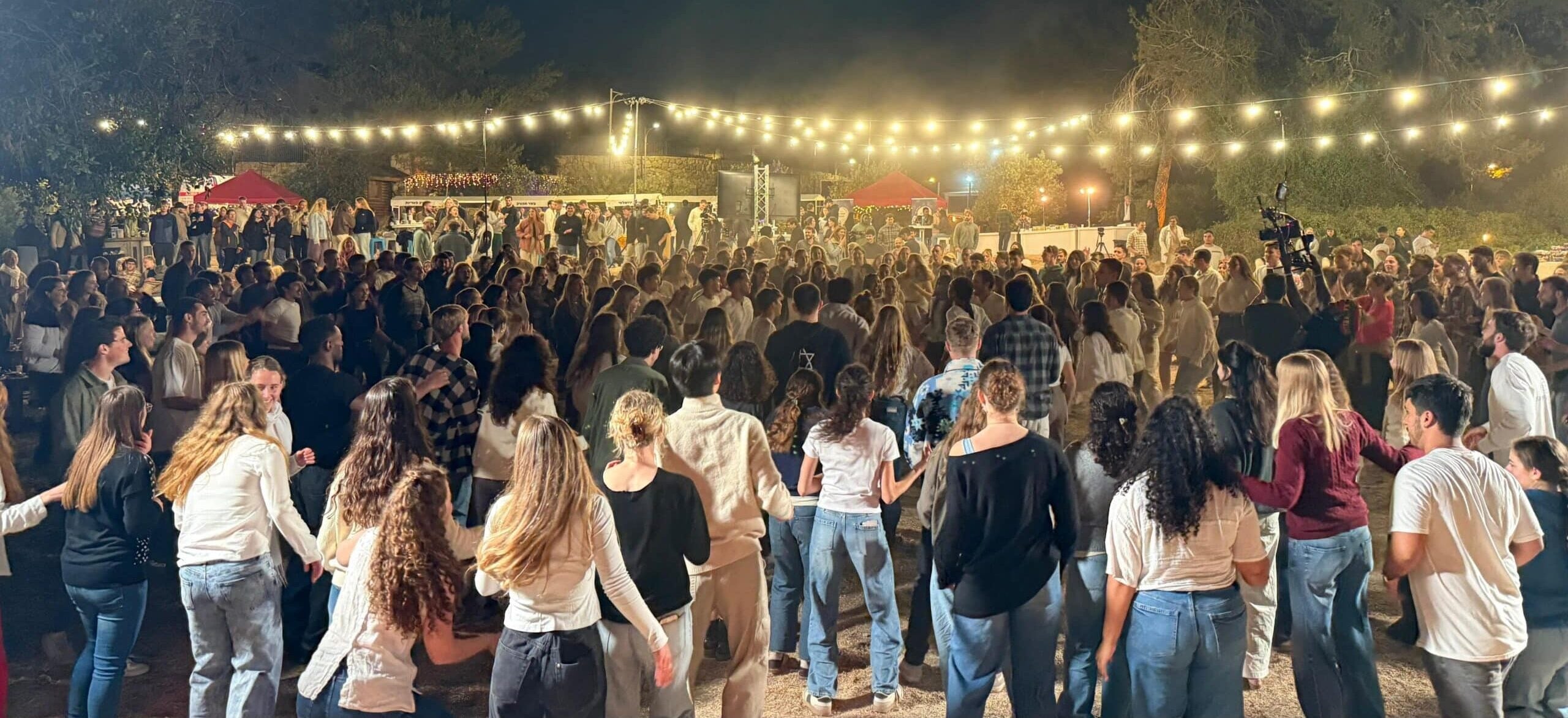 Large crowd dancing at an outdoor night event under string lights with stalls in the background.
