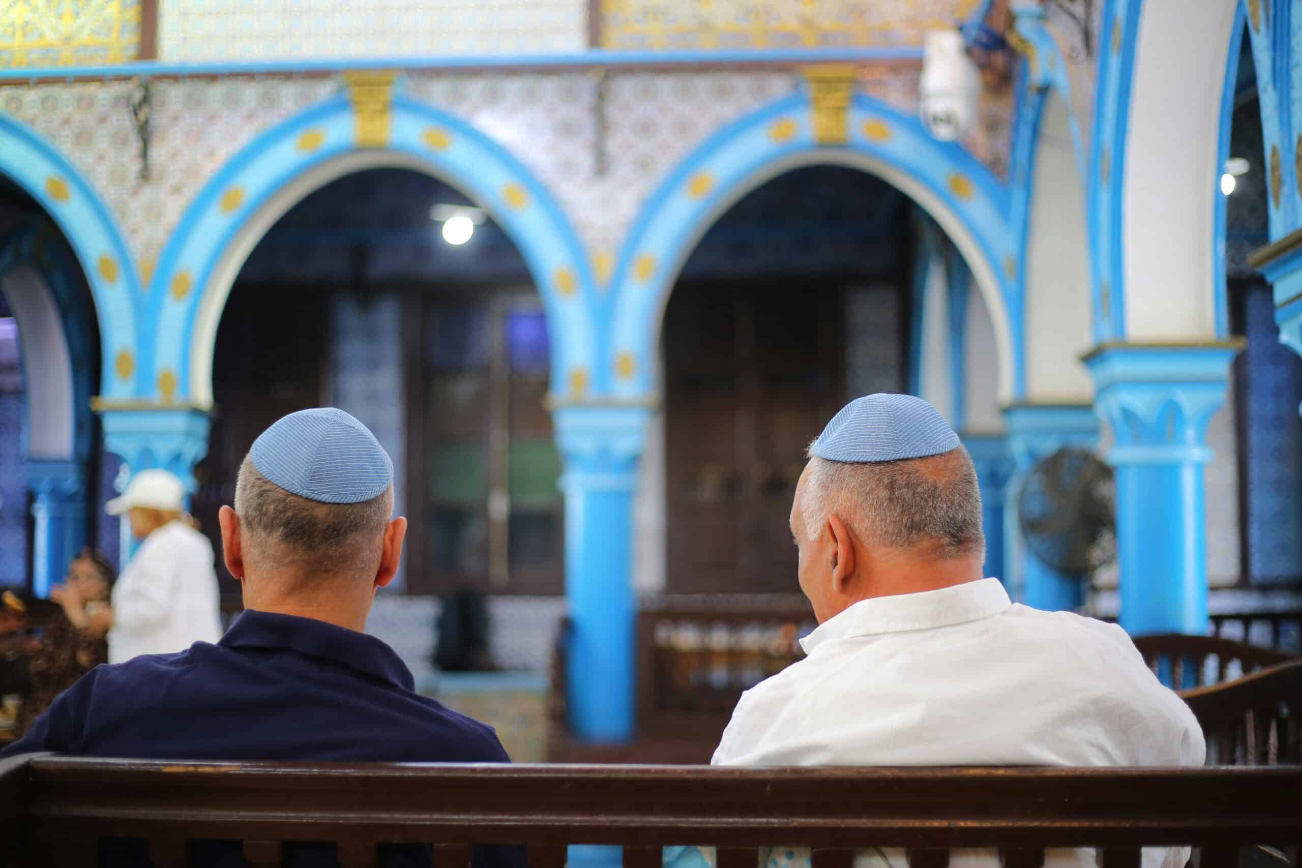 Two men wearing blue kippahs sit on a bench, facing a blue-arched courtyard interior with columns and decorative tiles.