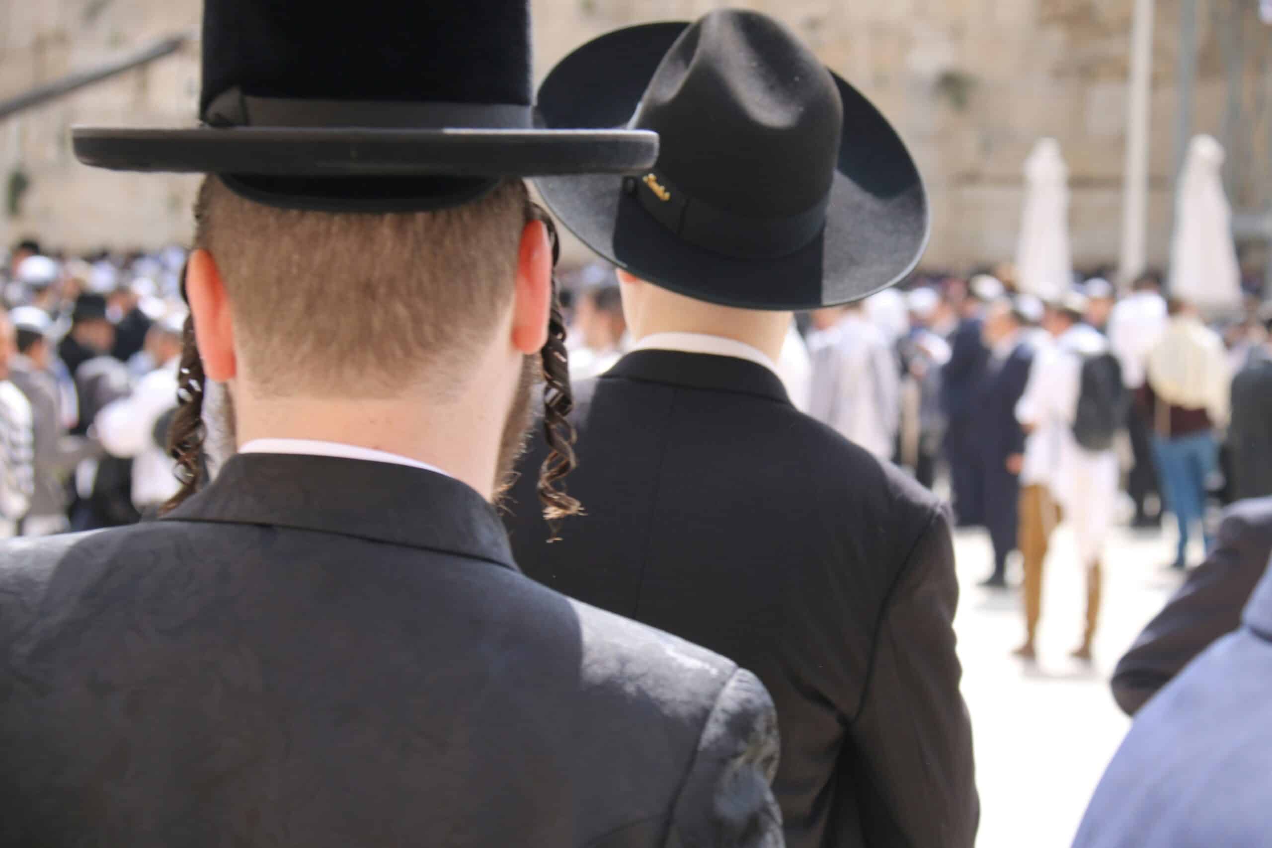 Back view of two men in black hats and suits among a busy outdoor crowd, seen from behind during a daytime gathering.