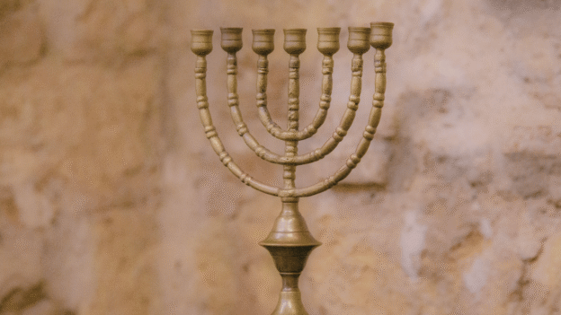 Bronze menorah with multiple curved branches on a dark wooden surface, set against a warm stone wall backdrop.