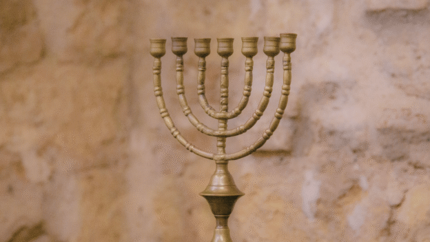 Bronze menorah with multiple curved branches on a dark wooden surface, set against a warm stone wall backdrop.