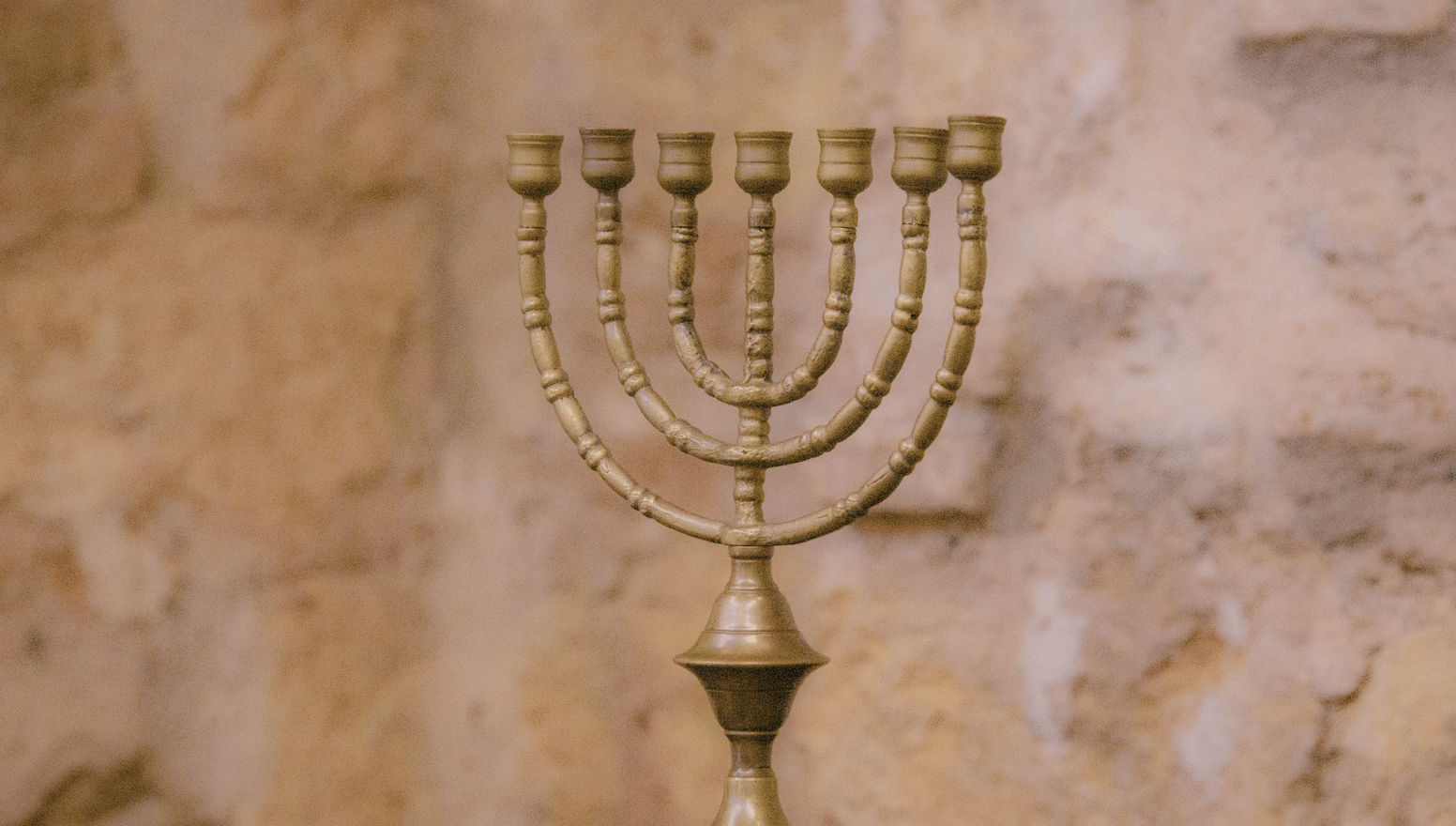 Bronze menorah with multiple curved branches on a dark wooden surface, set against a warm stone wall backdrop.