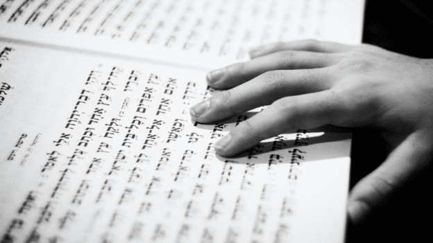 Close-up of a hand resting on a page of Hebrew text in a book.