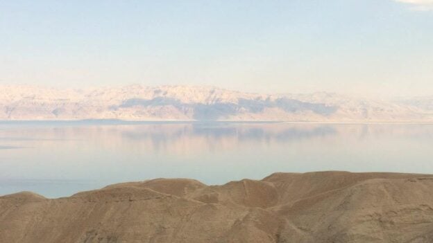 Desert hills in the foreground with a calm reflective lake and pale snow‑capped mountains in the distance under a blue sky.