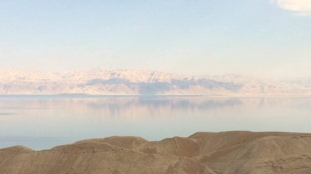 Desert hills in the foreground with a calm reflective lake and pale snow‑capped mountains in the distance under a blue sky.