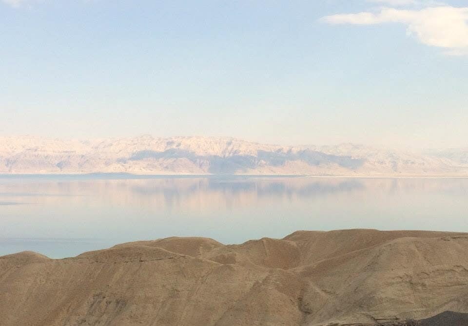Desert hills in the foreground with a calm reflective lake and pale snow‑capped mountains in the distance under a blue sky.