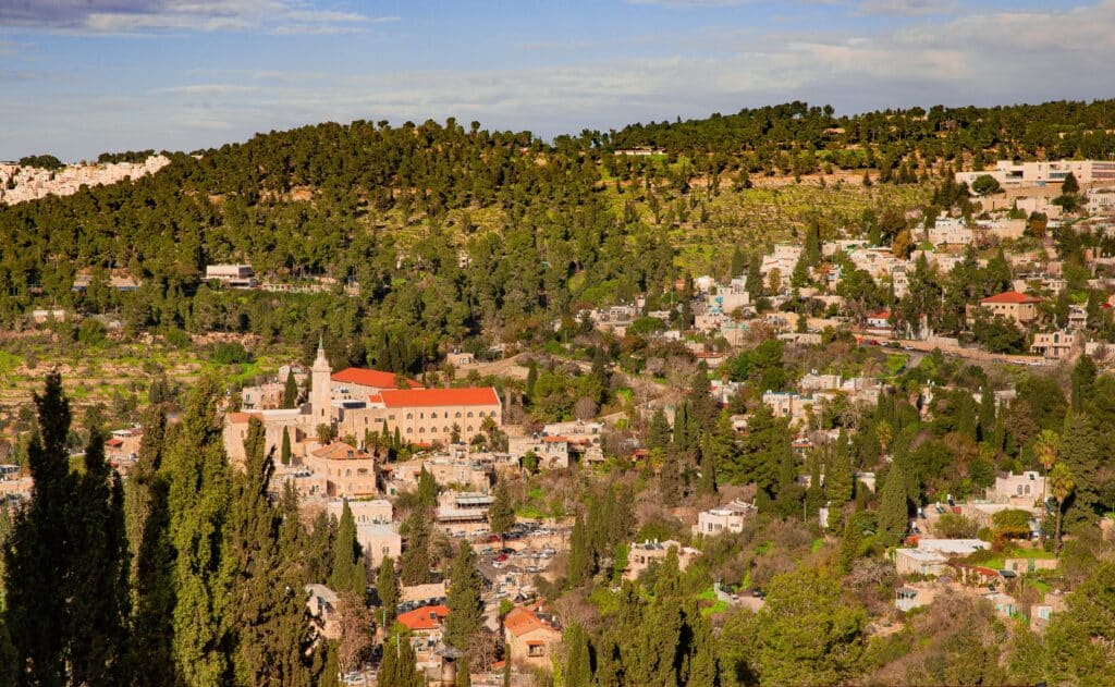 Hilly town with many trees and beige buildings; a church with a red roof sits in the foreground.