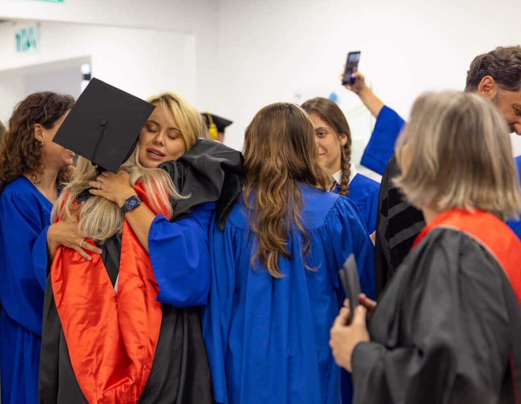 Graduate in a blue gown hugs a friend wearing a black cap during a graduation ceremony.