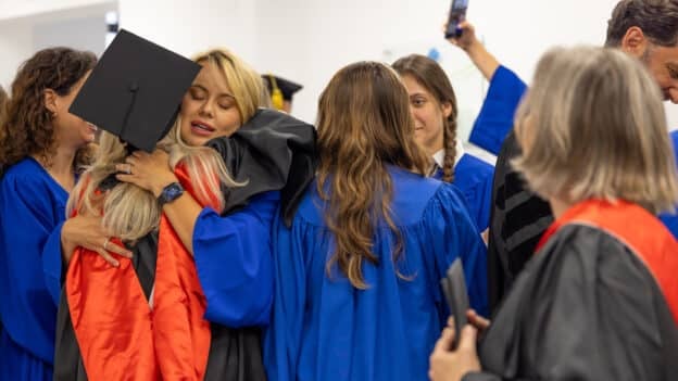 Graduate in a blue gown hugs a friend wearing a black cap during a graduation ceremony.