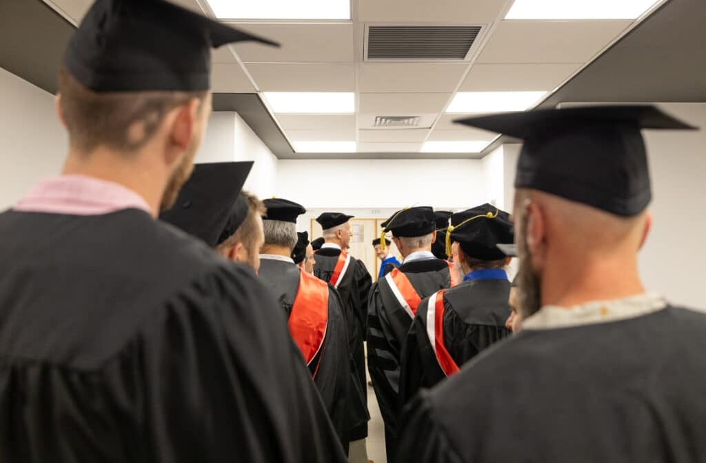 Group of graduates in black gowns and mortarboards gathered in a hallway, preparing for a ceremony.