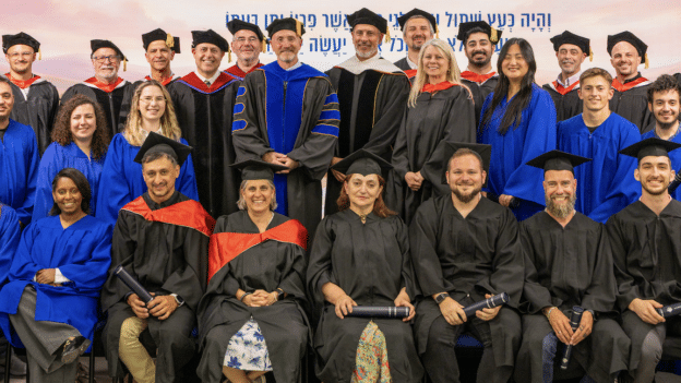 Group of graduates and faculty posing for a graduation ceremony photo; many wear blue robes, some in black gowns, holding diplomas in front row.