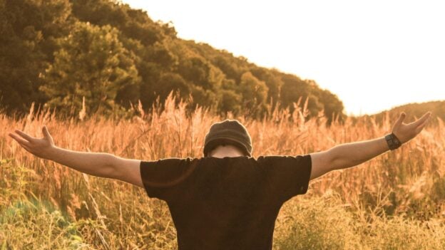 Person standing in a tall grassy field with arms outstretched at sunset, seen from behind.