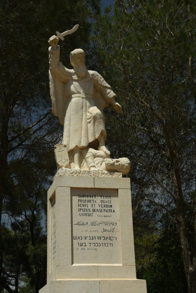 Stone statue of a robed bearded man raising a curved blade aloft, set on a stepped pedestal with multilingual inscriptions, surrounded by trees.
