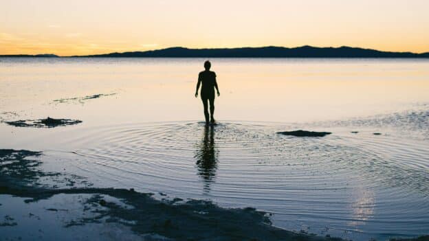 Silhouette of a person standing in shallow water at sunset, with calm sea and distant mountain range on the horizon.