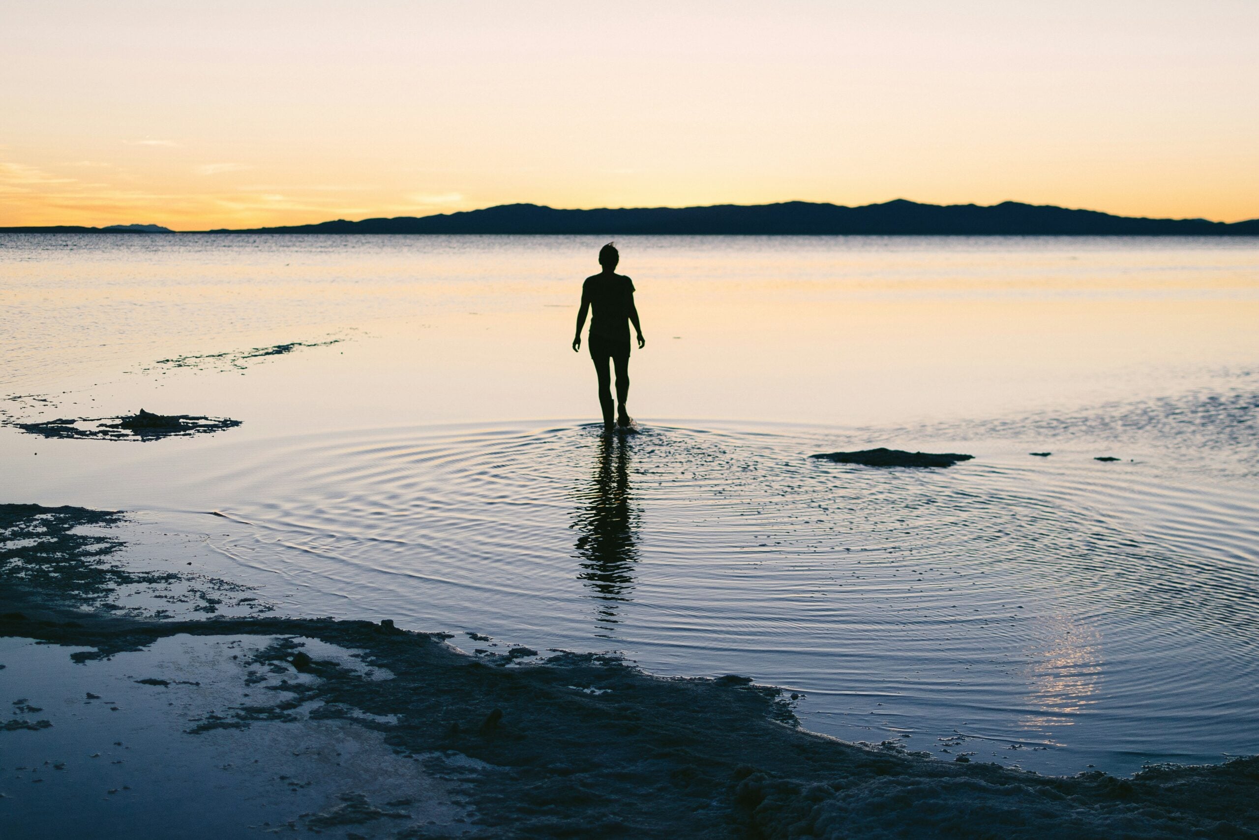 Silhouette of a person standing in shallow water at sunset, with calm sea and distant mountain range on the horizon.