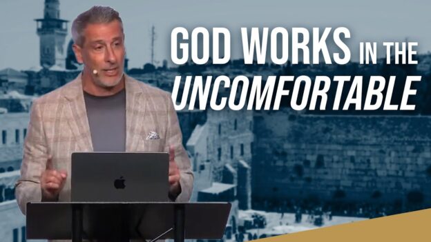 Speaker at a podium with a laptop, delivering a talk about 'God Works in the Uncomfortable' against a blue cityscape backdrop.