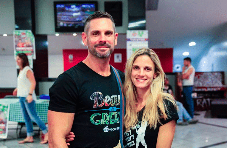 Smiling man and woman stand side by side in a bright indoor space, wearing casual black t-shirts, with posters and people in the background.
